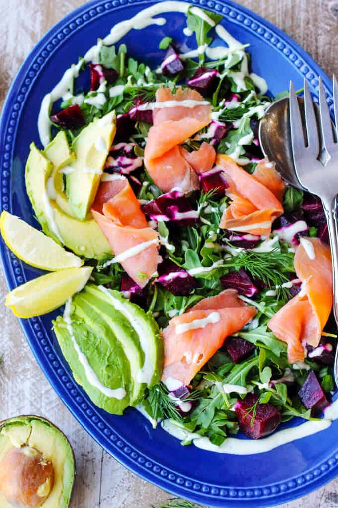 Smoked Salmon and Roasted Beets Salad with avocado, lemon slices a Goat Cheese Dressing on a blue plate with fork and spoon, half avocado on a side