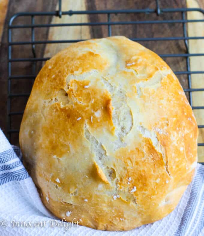This Homemade Dutch Oven Crusty Bread tastes like the fresh bread straight from the best bakery. But it comes from your oven and it only takes 10 minutes of prep time. There is no kneading involved! Homemade Dutch Oven Crusty Bread on a sheet rack with kitchen towel