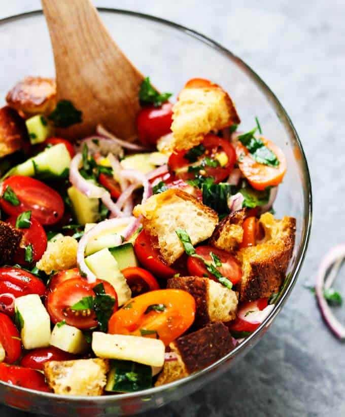 This popular Italian salad is a perfect addition to any summer meal or BBQ. Crusty bread that's been infused with garlic butter makes this it irresistible. Next time if you're looking for a perfect side dish try this amazing Summer Panzanella with Garlic Butter Bread. Italian Bread Salad with tomatoes, onions and cucumbers in a bowl with wooden spoon.