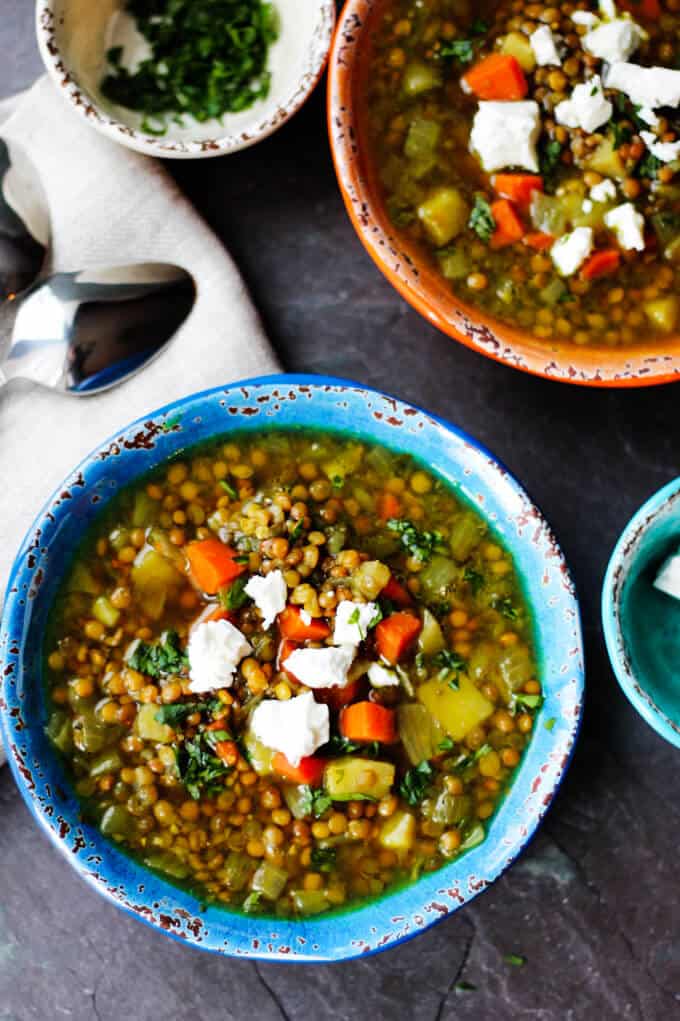 Two bowls: blue and orange of Fakes (Greek Lentil Soup) with feta with spoon and chopped parsley on a side. 