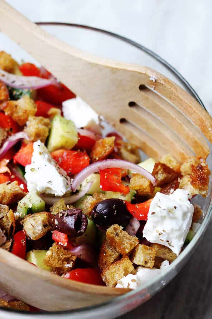 Greek Panzanella Salad combines traditional Greek salad flavors, like Kalamata olives, and tangy feta with bread cubes toasted in tasty garlicky vinaigrette. Greek Panzanella Salad in a bowl with wooden spoon and fork, combines traditional Greek salad flavors, like Kalamata olives, and tangy feta with bread cubes toasted in tasty garlicky vinaigrette.