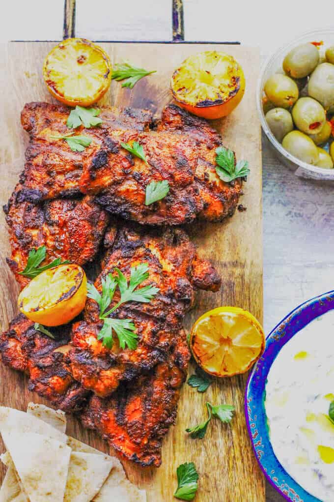 Grilled Chicken Shawarma on a cutting board with grilled lemons, olives and yogurt sauce on a side 