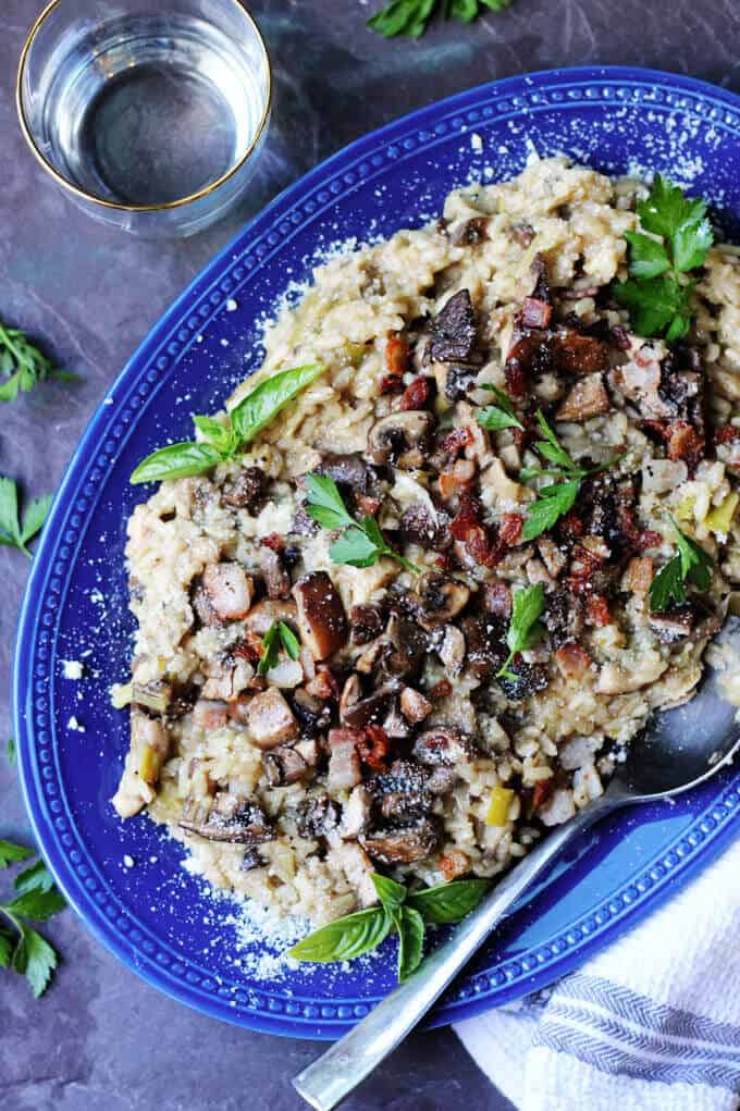 Wild Mushroom Risotto with Bacon and Leeks is exceptionally flavorful and delicious. Every bite introduces a combination of different flavors and textures. Mushroom Risotto With Bacon on a blue plate with glass of water on a side