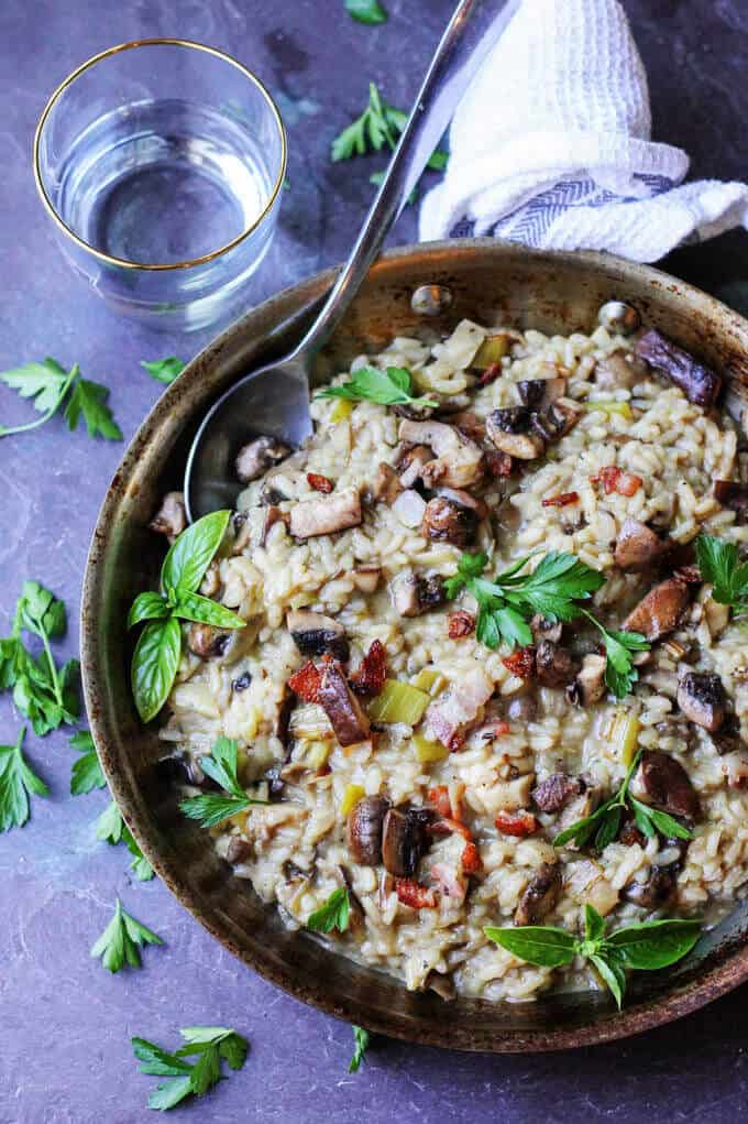 Wild Mushroom Risotto with Bacon and Leeks is exceptionally flavorful and delicious. Every bite introduces a combination of different flavors and textures. Wild Mushroom Risotto with Bacon and Leeks in a skillet with spoon and water on a side