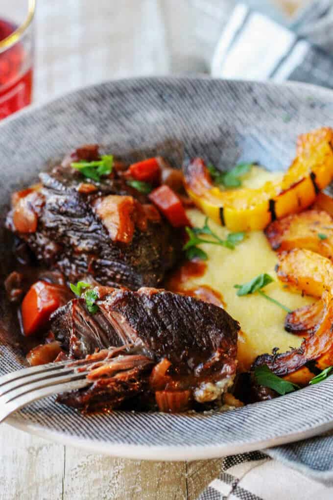Red Wine Braised Short Ribs with Parmesan Polenta and Roasted Delicata Squash, is a super flavorful dinner idea that will make you feel warm and cozy on a cold, winter night. Red wine braised short ribs with parmesan polenta and delicata squash on a plate with a fork