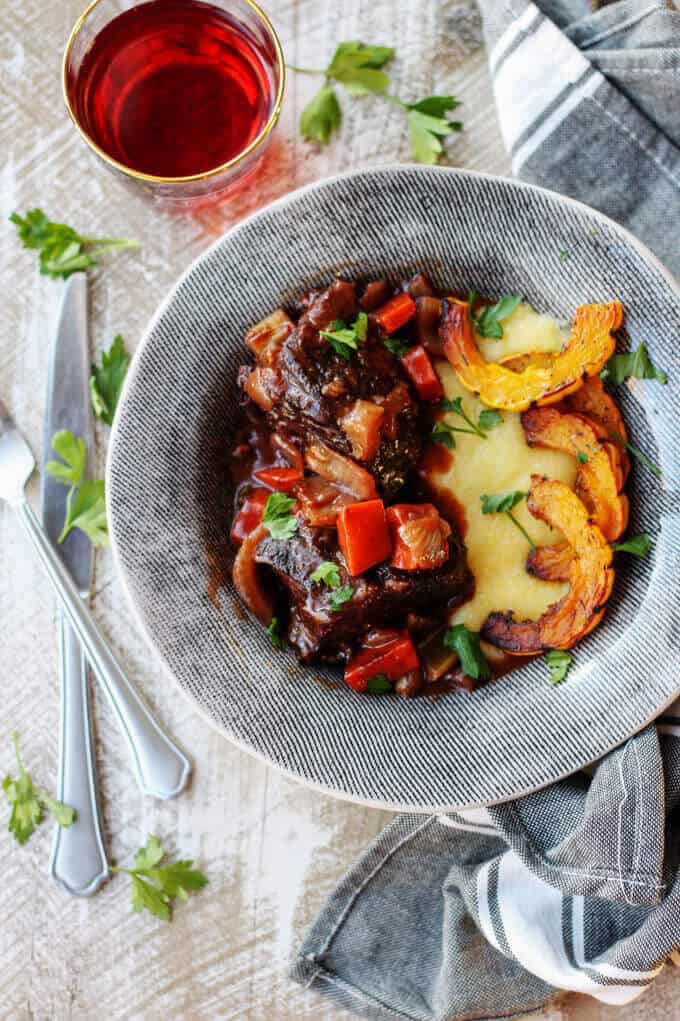 Red Wine Braised Short Ribs with Parmesan Polenta and Roasted Delicata Squash, is a super flavorful dinner idea that will make you feel warm and cozy on a cold, winter night. Red wine braised short ribs with polenta and squash in a grey plate on wooden background with drink on a side