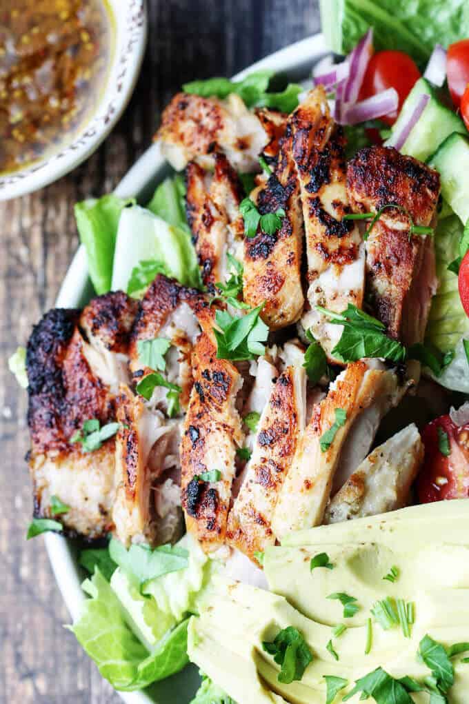Garlic Lemon Chicken Salad with tomatoes, cucumbers, onions, lettuce, avocado on a plate; half avocado on a side; dressing in a bowl with spoon on a side; frozen garlic and onion packets.