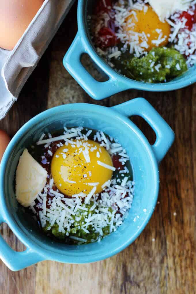 These Baked Eggs with Tomatoes and Pesto can be added to your ketogenic breakfast menu if you are looking for low carb recipes. It's super quick, delicious, naturally low in carbs and packed full of healthy fats. cracked eggs in ramekins over tomatoes, topped with pesto, butter and parmesan cheese