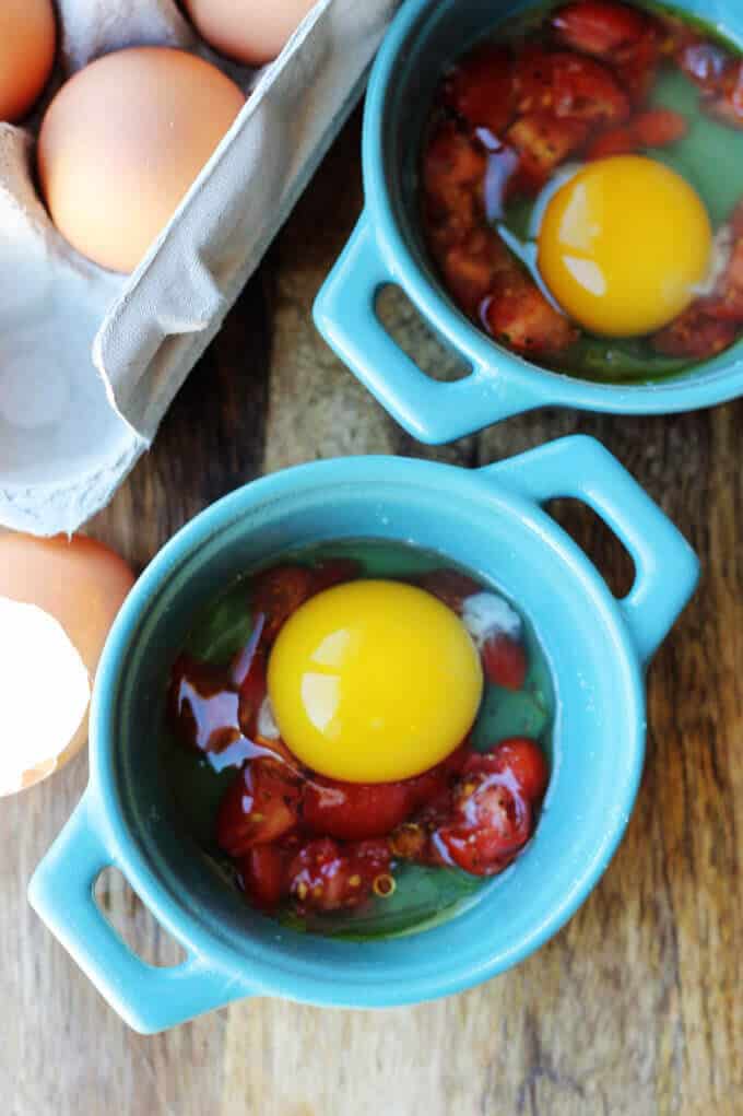 These Baked Eggs with Tomatoes and Pesto can be added to your ketogenic breakfast menu if you are looking for low carb recipes. It's super quick, delicious, naturally low in carbs and packed full of healthy fats. cracked eggs over tomatoes in blue ramekins,