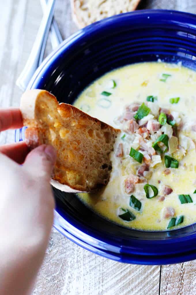 This Ham and Cheese Soup with White Wine and Gruyere is yet another recipe to show you how easily you can repurpose your leftover ham. This soup is rich and decadent, and it will keep your loved ones coming back for seconds...guaranteed. ham and cheese soup with scallion in blue bowl with hand dunking the bread in the soup