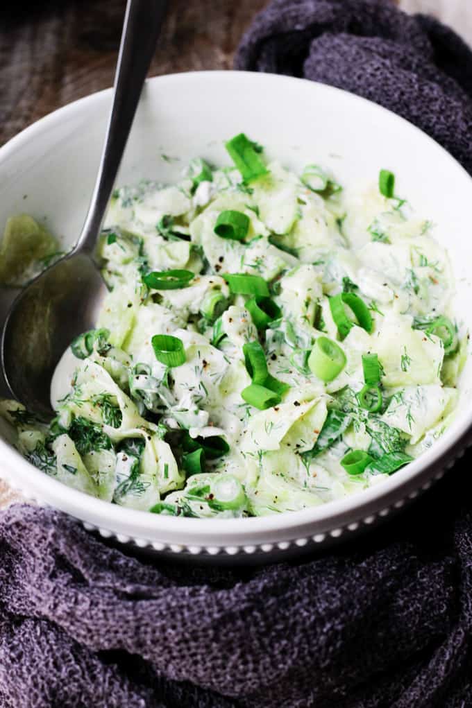 With summer around the corner, so comes the need for nice, refreshing dishes. This Polish Cucumber Salad (Mizeria) ought to find its way to your summer dishes repertoire. Simple ingredients like cucumber, dill, green onions and sour cream is all you need. Polish cucumber salad - mizeria in a bowl with spoon in it and towel around it