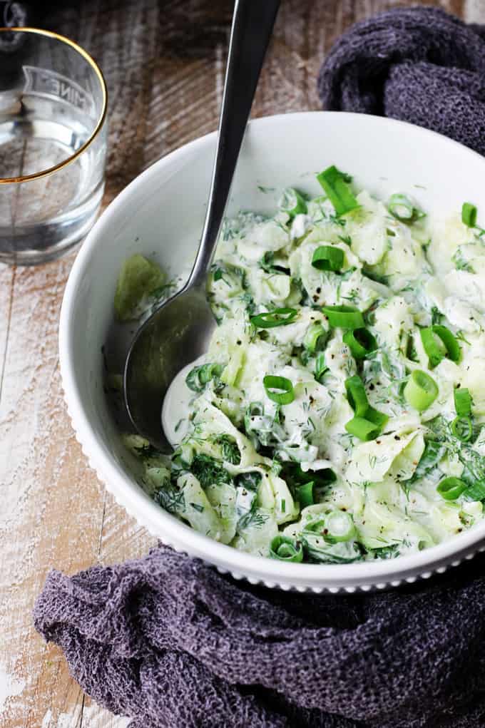 With summer around the corner, so comes the need for nice, refreshing dishes. This Polish Cucumber Salad (Mizeria) ought to find its way to your summer dishes repertoire. Simple ingredients like cucumber, dill, green onions and sour cream is all you need. Polish cucumber salad in a white bowl with spoon in and towel around; water in a back