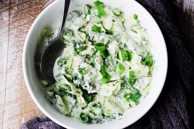 With summer around the corner, so comes the need for nice, refreshing dishes. This Polish Cucumber Salad (Mizeria) ought to find its way to your summer dishes repertoire. Simple ingredients like cucumber, dill, green onions and sour cream is all you need. Polish cucumber salad in a white bowl with spoon; overhead shot, horizontal