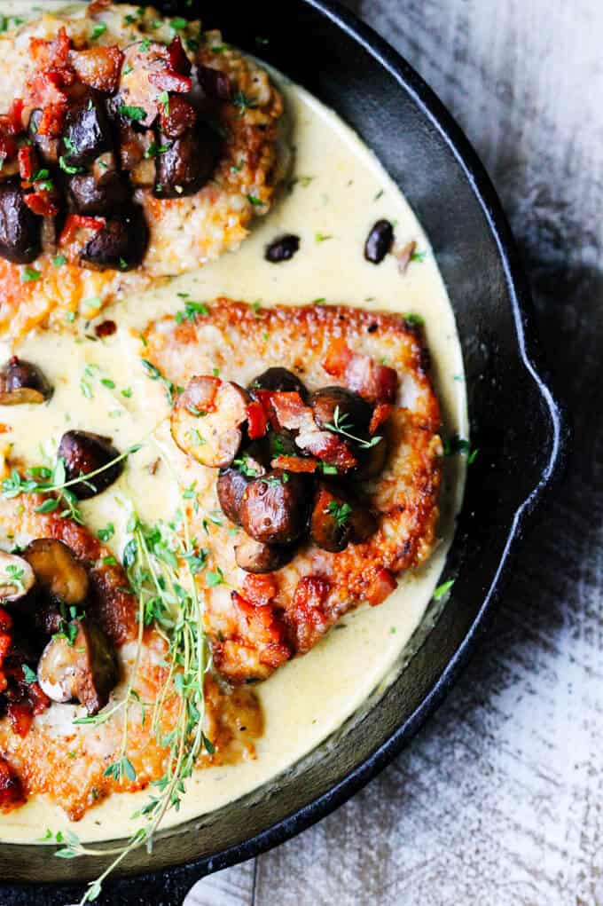 Pork chops in white wine cream sauce with mushrooms and bacon on top in a pan, overhead shot