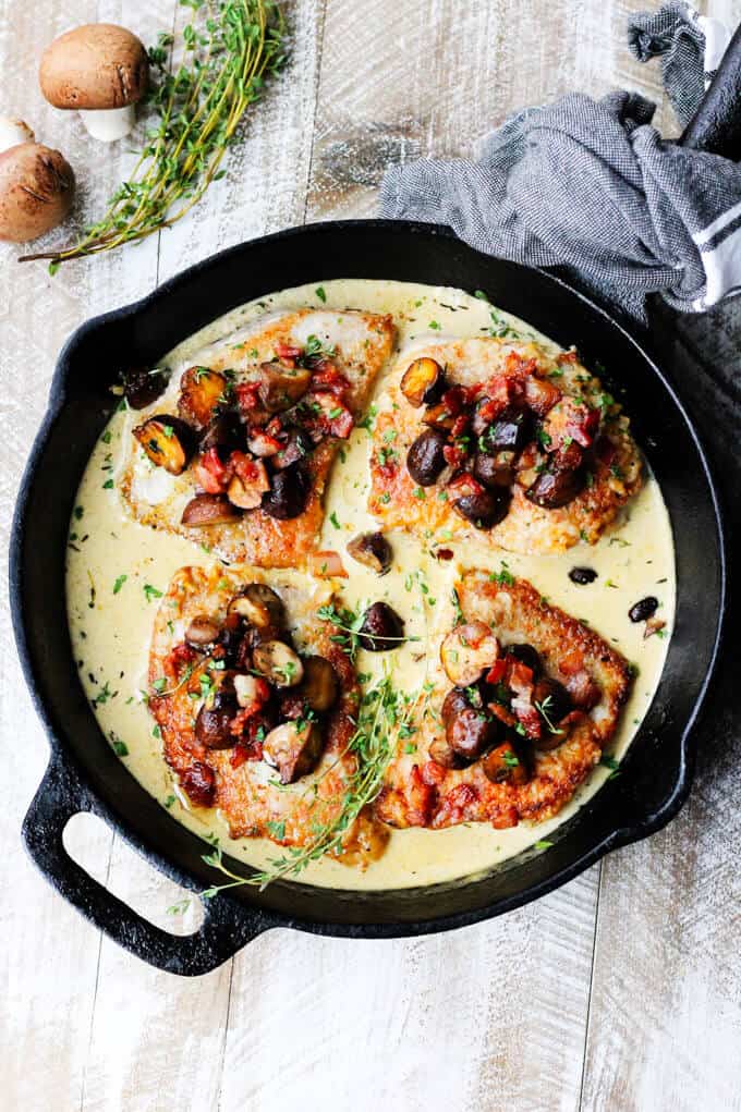 Pork chops in white wine creams sauce in a pan, diagonal overhead shot with mushrooms and thyme on a side