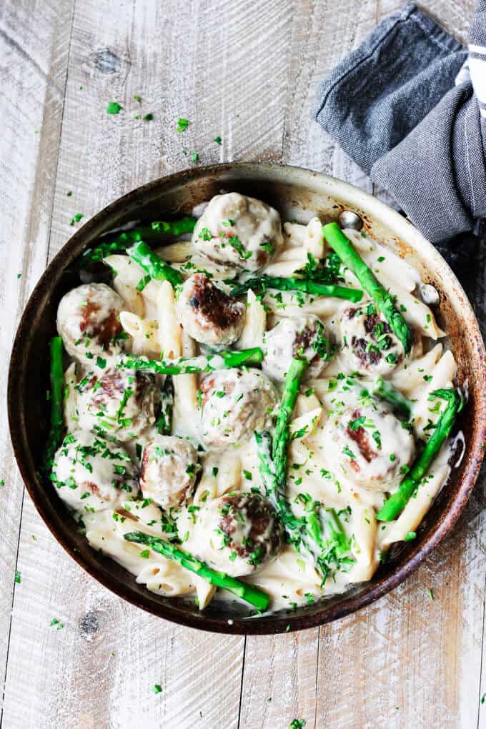 This Turkey Meatballs Alfredo Pasta with Asparagus is a perfect midweek dinner, for nights when you're short on time but still want to make your family a hearty meal. It comes together quickly and incorporates a store-bought Alfredo sauce shortcut. Turkey meatballs with asparagus in Alfredo sauce in a pan with kitchen towel; overhead shot on a board