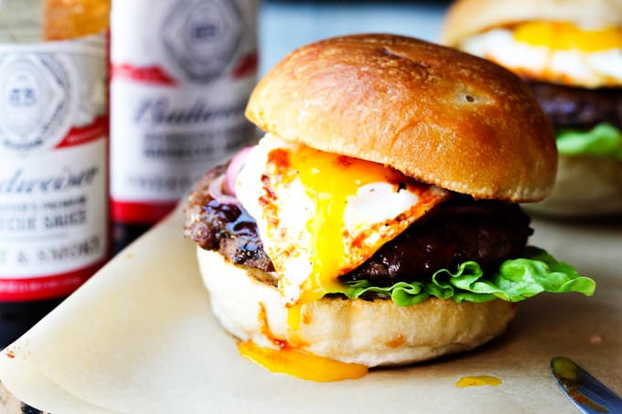 Smoky BBQ Burger with Garlic Aioli & Fried Egg is a perfect summer meal that uses Budweiser Sweet & Smoky BBQ sauce, beef patties, fried eggs, pickled onions and garlic aioli. Smoky BBQ burger with runny egg yolk and sauces in the back