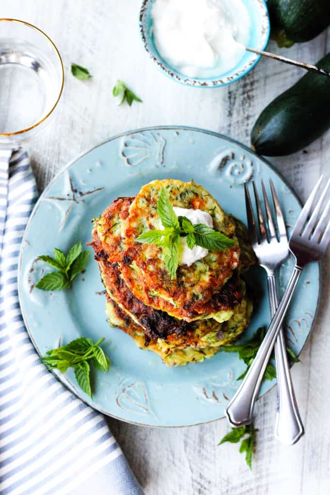 Overhead photo of Greek zucchini fritters on a blue plate with yogurt on a side and a glass of water