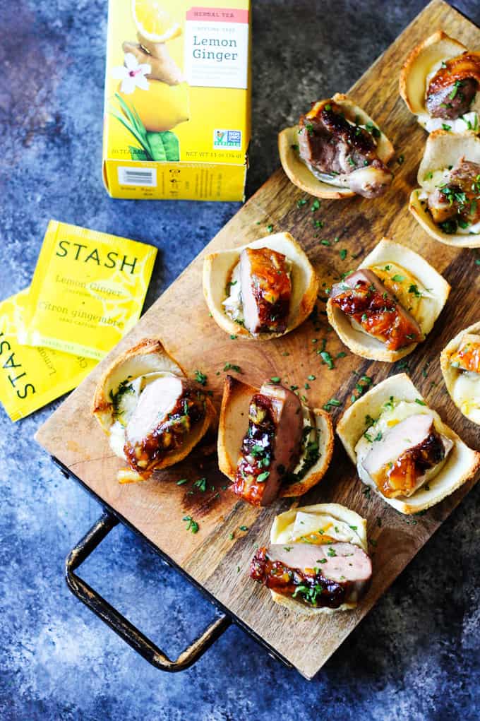 Tea Infused Orange Duck Bites with Brie are a perfect appetizer for Easter or any other Holidays. They are full of flavor with Stash Lemon Ginger Tea infused duck breast, seared to perfection, smothered with Lemon Ginger Tea Orange Glaze and topped on the melted brie toasts for the ultimate bite. Tea Infused Suck Bites with Brie on a serving board with tea bags on a side