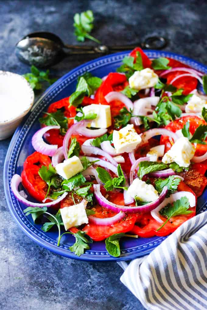 This Tomato Feta Salad is as simple as it gets, and it is super delicious! When ripe, juicy tomatoes are in season you'll want to eat it every day. It contains a few simple ingredients and it comes together in no time at all. Tomato Feta Salad on a blue serving plate with spoon at the back and kitchen towel