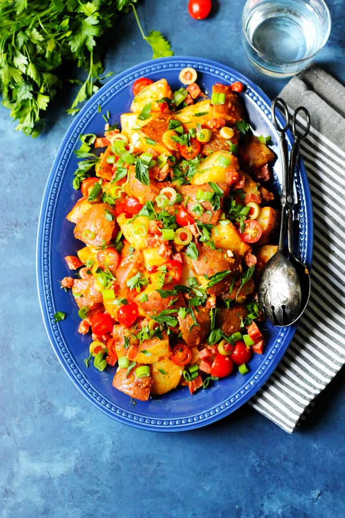 Spanish Potato Salad with Smoked Paprika and Chorizo on a blue serving dish with kitchen towel and glass of water on a side