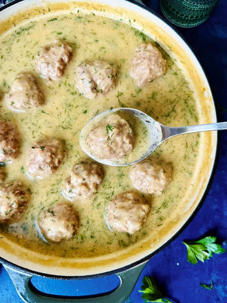 Boiled meatballs and sauce, in a pan, with one meatball lifted out on a spoon
