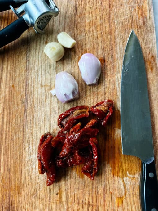 Shallot, garlic and sun-dried tomatoes on a chopping board with a knifr