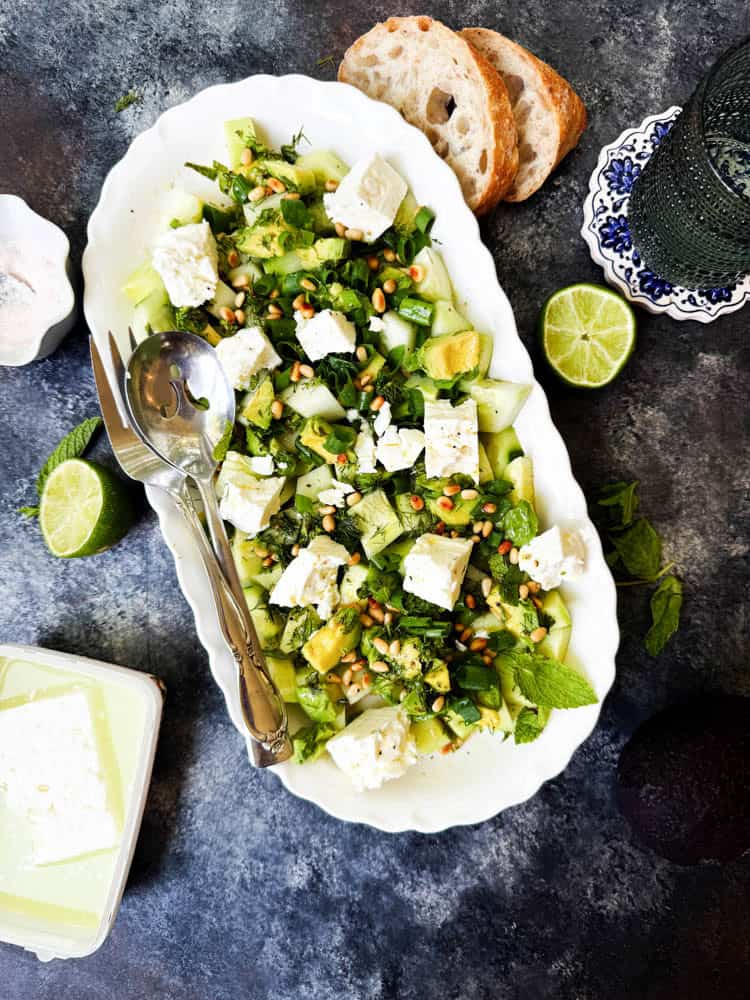 Overhead view of cucumber feta cheese salad on a white plate surrounded by lime, feta and two slices of bread